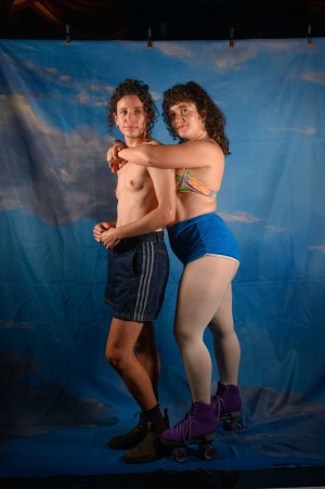 Grace, a brunette person wearing a bikini and blue shorts, puts her arms around Amit, who is topless. They both look into the distance.